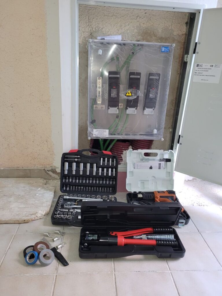 Un cuadro eléctrico abierto con cables gruesos de color verde conectados a interruptores y disyuntores. El cuadro tiene una cubierta transparente protectora. Delante del cuadro, en el suelo, hay varias cajas de herramientas abiertas que contienen diferentes tipos de llaves, destornilladores y enchufes. También hay un cortador de cables rojo, rollos de cinta adhesiva de varios colores y algunas piezas de conexión metálicas.
