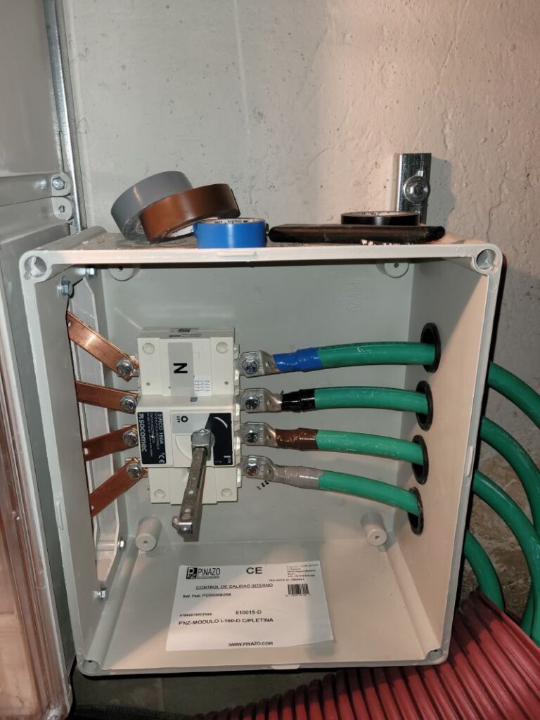 Un cuadro eléctrico abierto que muestra un interruptor y varios cables conectados. Los cables son gruesos y de color verde, conectados a terminales de cobre.