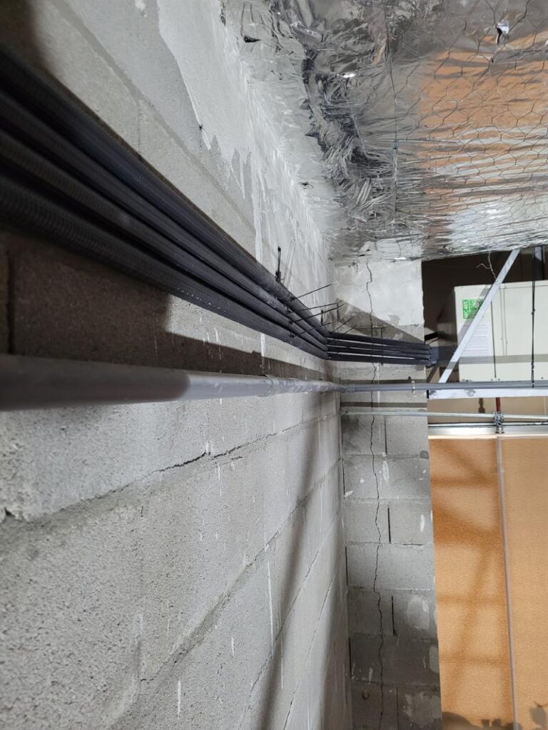 Varias tuberías negras corriendo a lo largo de una pared de concreto con aislamiento plateado en el techo. Las tuberías están bien organizadas y sujetas a la pared.