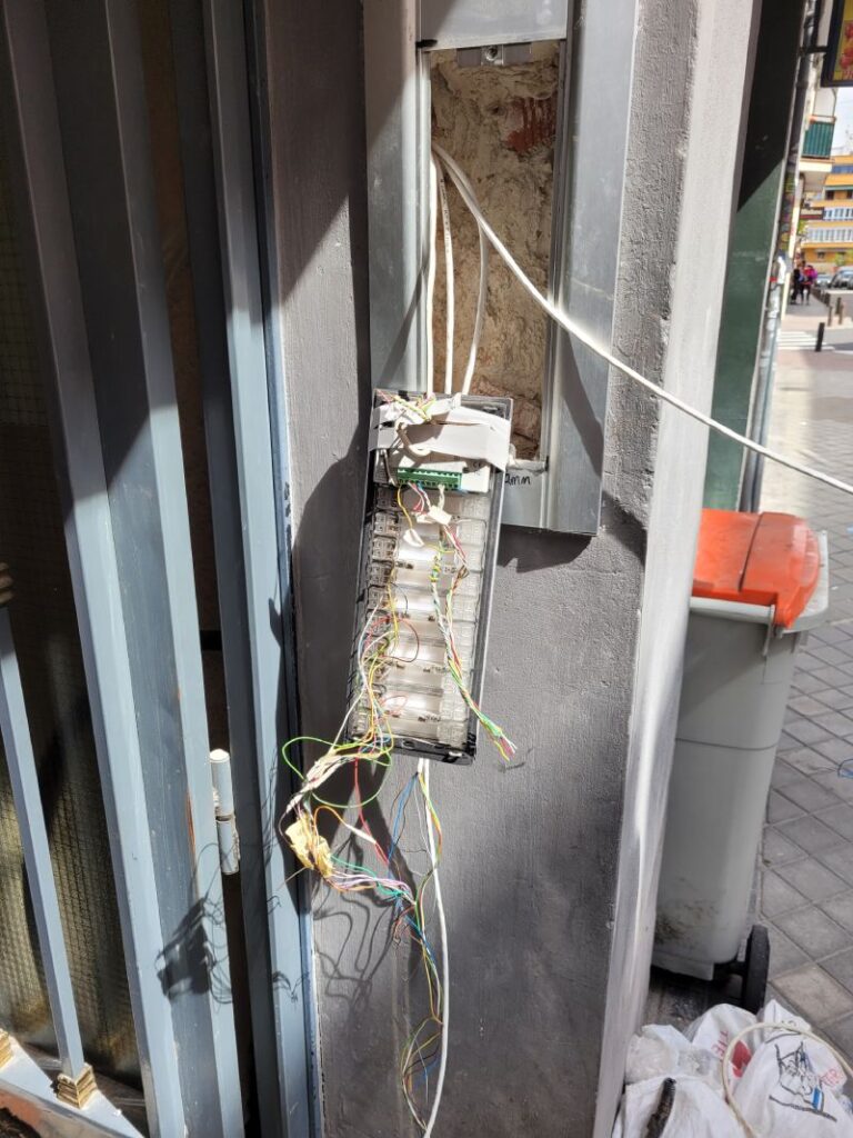 Un panel de control con cables expuestos colgando fuera de una caja en la pared exterior de un edificio. Los cables están desorganizados y hay una puerta abierta que revela más cables dentro.
