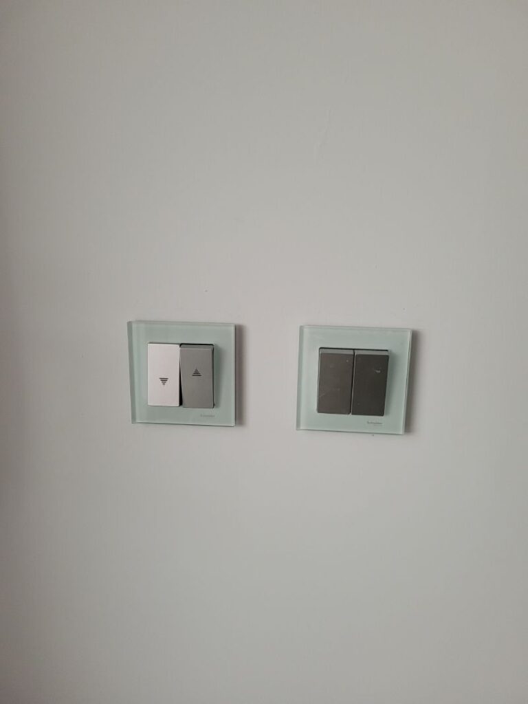 Dos interruptores de luz en una pared blanca. El interruptor de la izquierda tiene un símbolo de triángulo, indicando una función de subida y bajada, mientras que el de la derecha tiene dos interruptores estándar.