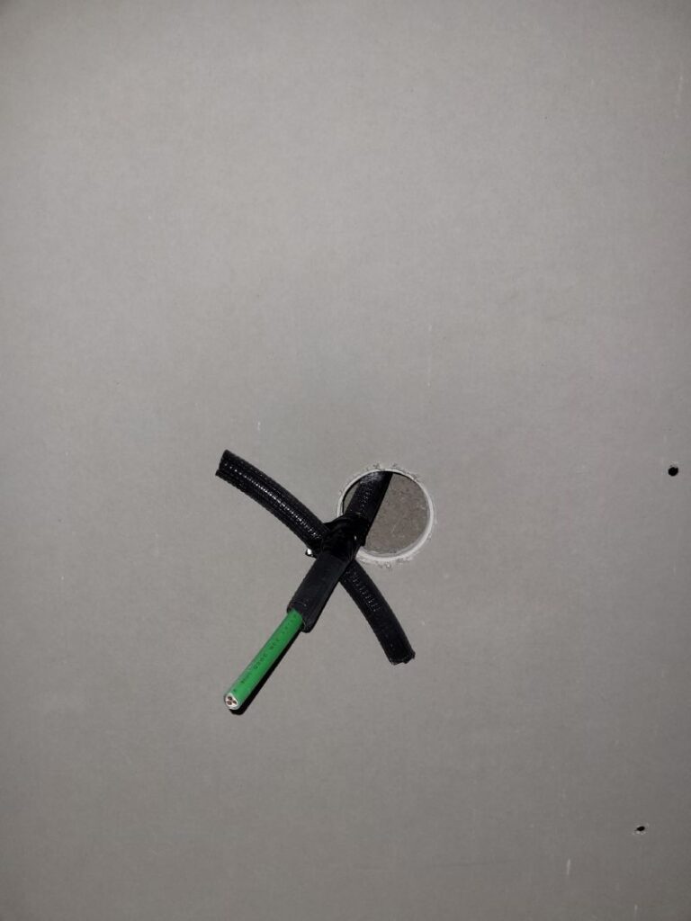 Cable verde y negro que sale de un agujero redondo en una superficie gris, probablemente una pared o techo de yeso